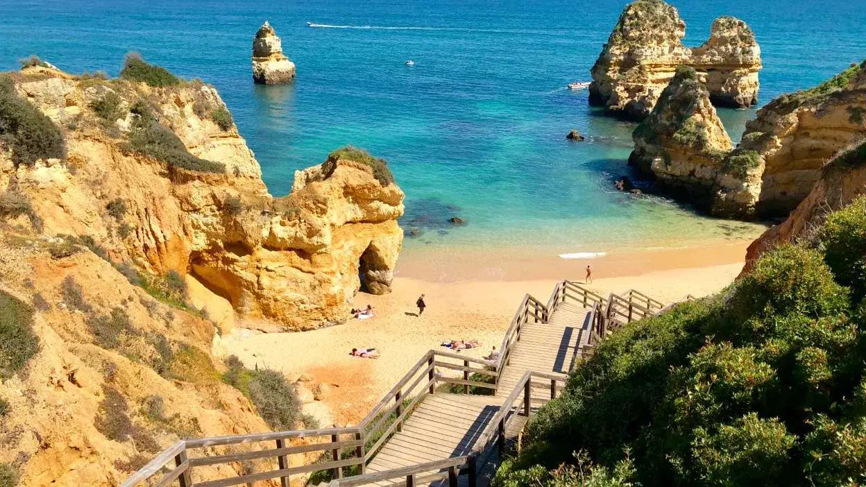 Escadaria de madeira a descer para praia com rochas e mar azul em Portimão, Algarve.