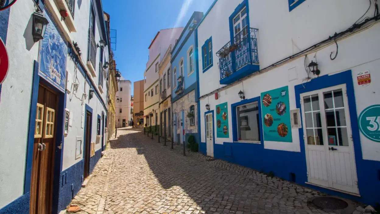 Rua estreita com calçada de pedra e casas coloridas em Portimão, Algarve, Portugal, sob céu azul.