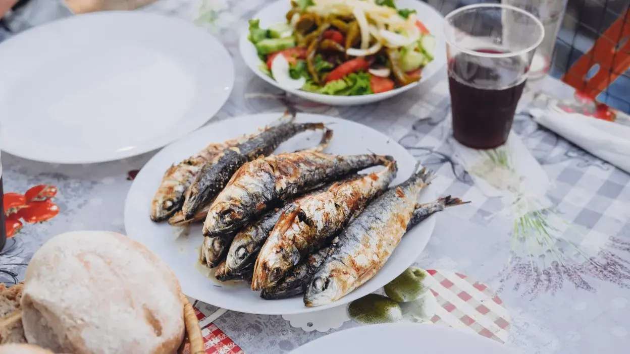Prato com sardinhas grelhadas, pão, salada e bebida numa mesa com toalha decorada.