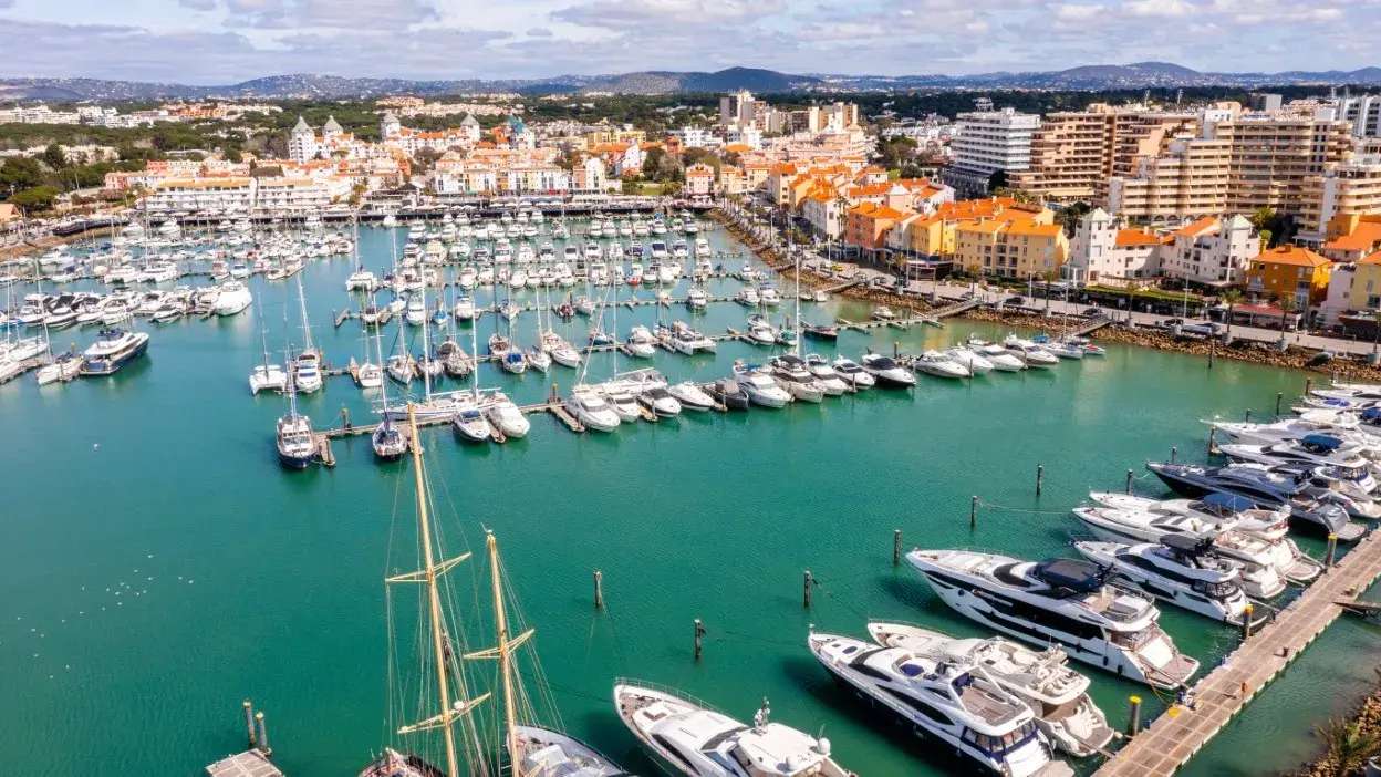 Vista aérea da marina de Vilamoura com vários iates atracados e edifícios ao fundo.