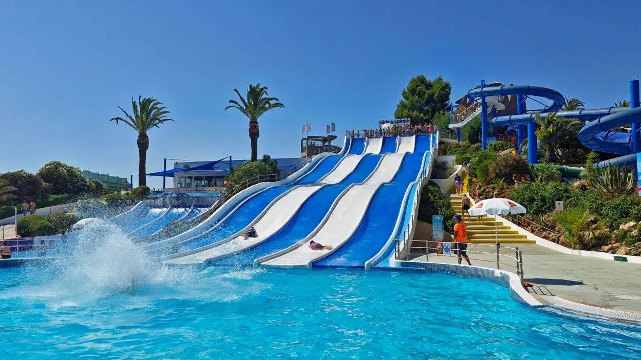 Escorregas azuis e brancos num parque aquático com piscina e palmeiras ao fundo.