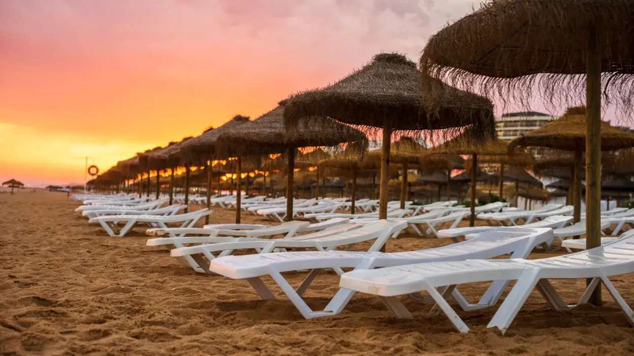 Fileira de espreguiçadeiras brancas e chapéus de palha numa praia ao pôr do sol
