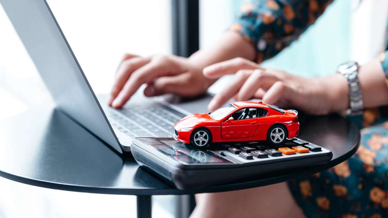 Senhora sentada a mexer no computador, com uma calculadora ao lado e um carro vermelho de brincar em cima da calculadora