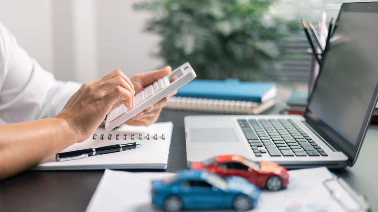 pessoa a fazer contas na calculadora rodeada de dois carrinhos de brincar, cadernos e um portátil