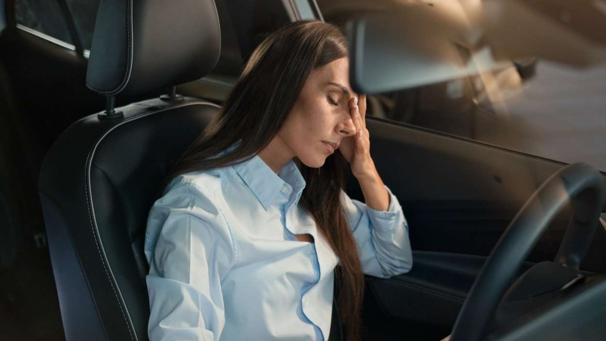 mulher sentada dentro do carro, visivelmente cansada, com olhos fechados e cabeça apoiada na mão